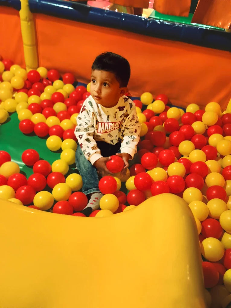 A young child sits in a ball pit, holding a red ball.