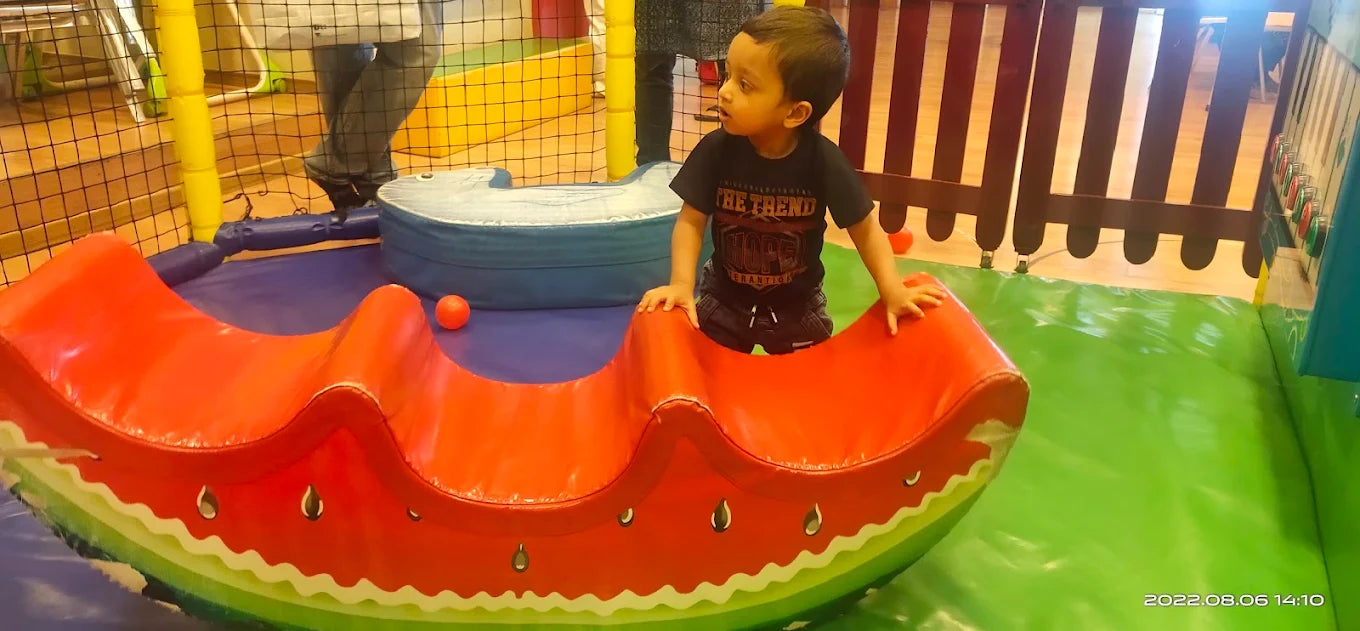 A young boy plays on a watermelon-shaped rocker in a colorful indoor playground.