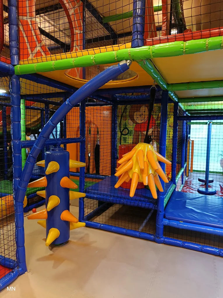 A colorful indoor playground with soft blue and yellow structures, featuring a spiky blue pillar and a hanging orange plaything.