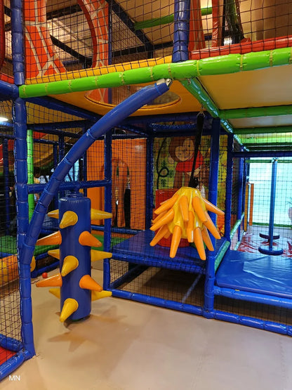 A colorful indoor playground with soft blue and yellow structures, featuring a spiky blue pillar and a hanging orange plaything.