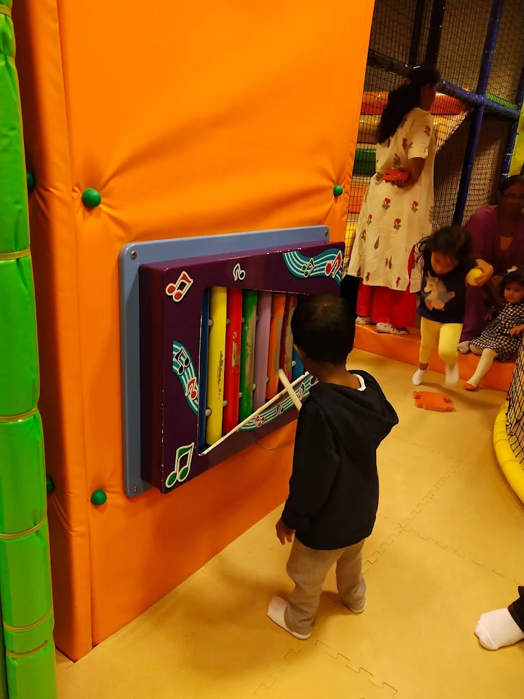 A young boy plays a xylophone in a colorful indoor playground. Other children and adults are visible in the background.