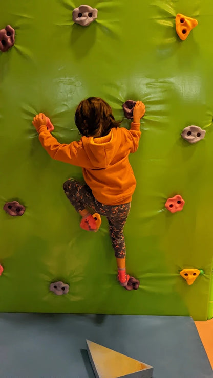 A young child climbs a green wall with colorful holds.