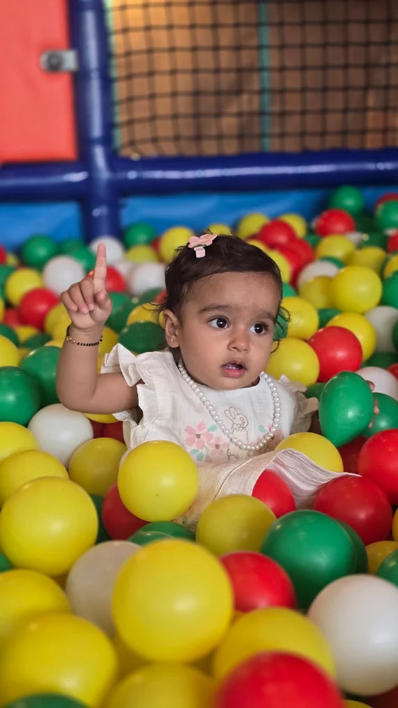 A baby girl sits in a ball pit, pointing her finger up. She is surrounded by colorful plastic balls.