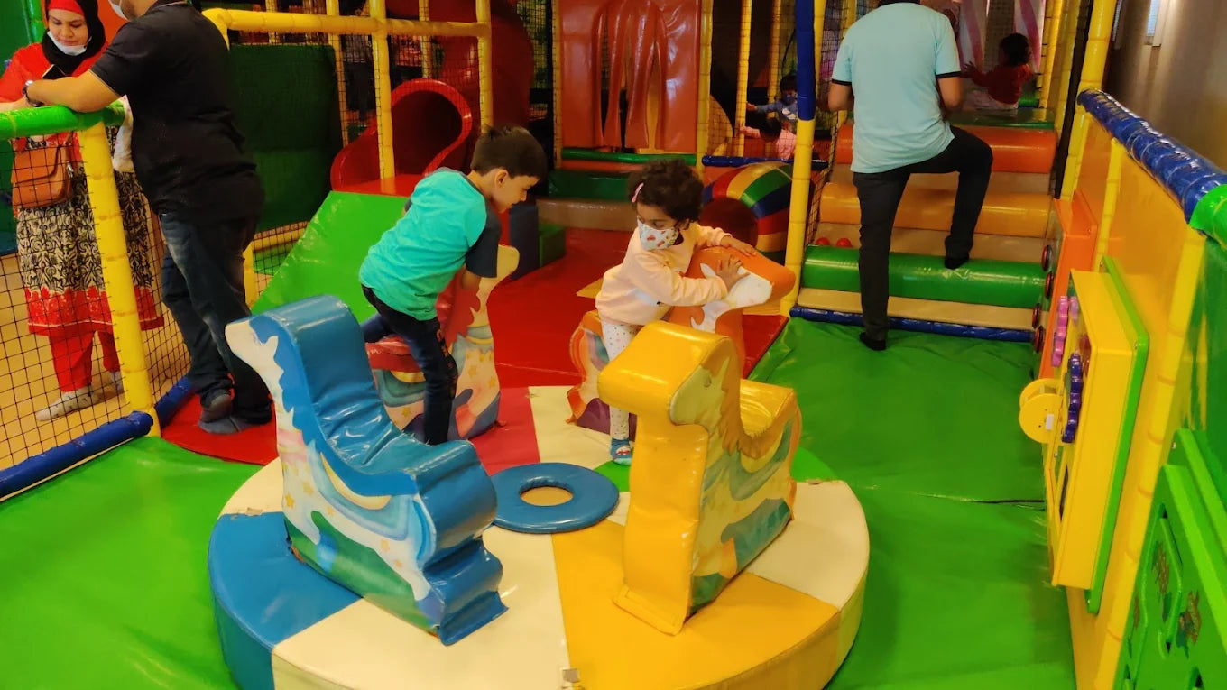 Children play on colorful rocking animals in a soft play area. Adults supervise in the background.