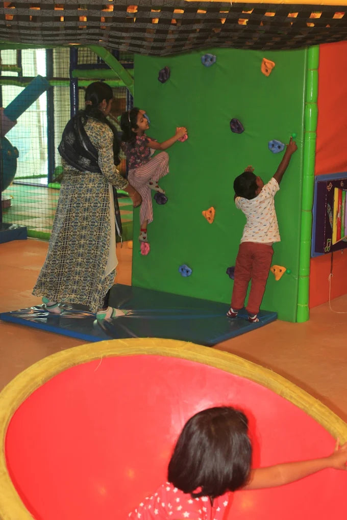 Children climb a green wall at an indoor playground while a woman supervises.