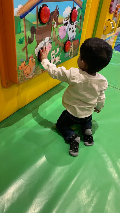 A young child kneels and plays with a farm-themed interactive wall in a brightly colored play area.