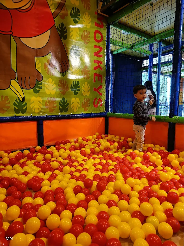 A young child stands in a ball pit filled with red and yellow balls, with a monkey-themed mural in the background.