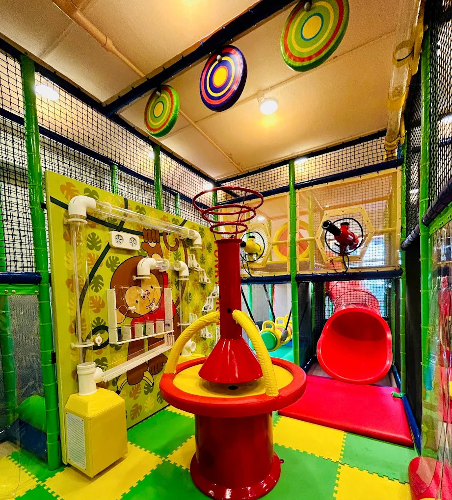 A colorful indoor playground with a monkey-themed activity wall, a ball-toss game, and a red tube slide for children.