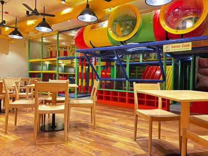 A brightly colored indoor playground with tables and chairs in the foreground. The playground features tubes, slides, and climbing structures for children.