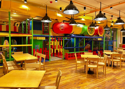 A colorful indoor playground with tables and chairs in the foreground. The playground features tubes, slides, and climbing structures for children.
