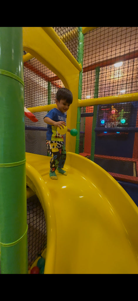 A young boy plays on a yellow slide in an indoor playground. He is holding two colorful balls.