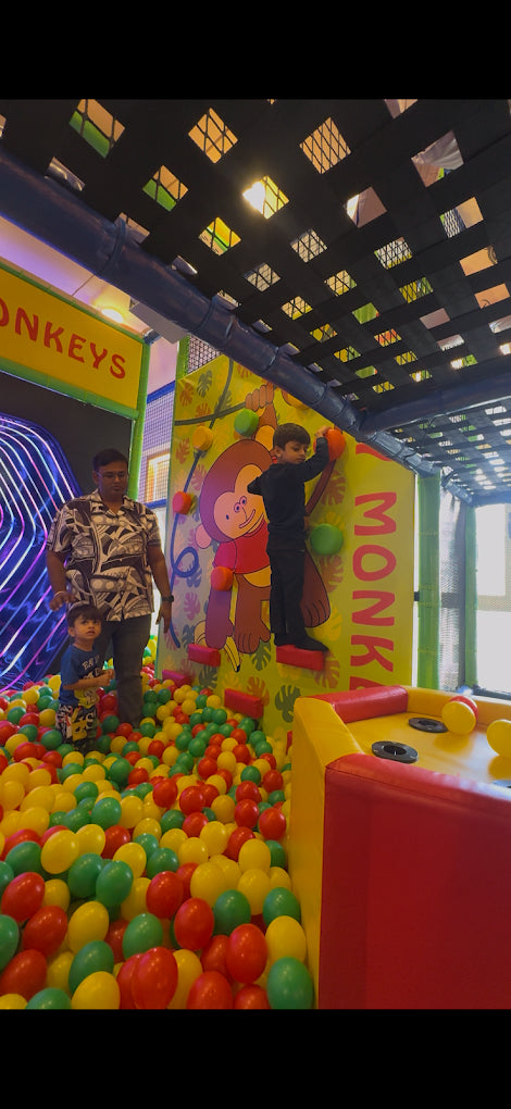 A young boy climbs a wall in a ball pit while a man and another young boy watch.