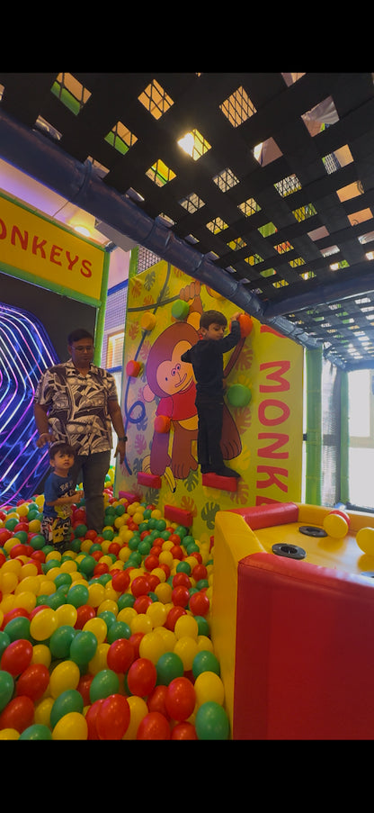 A young boy climbs a wall in a ball pit while a man and another young boy watch.