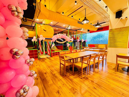 A brightly lit indoor playground with tables and chairs in the foreground, decorated with pink balloons.