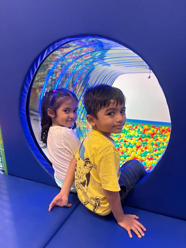 Two young children sit in a blue tunnel overlooking a ball pit.