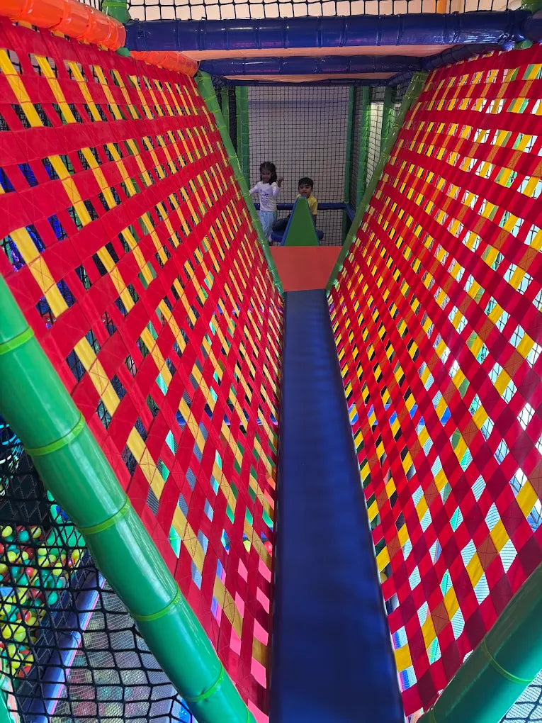 Two young children play in a colorful indoor playground with red, yellow, and blue netting.