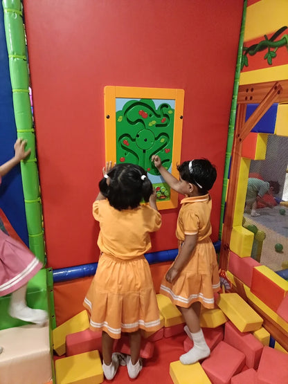 Two young children play with a maze toy on a red wall in a colorful indoor playground.