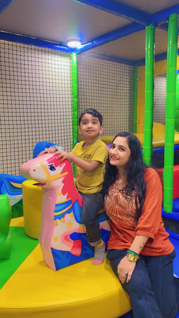 A woman and a young boy are playing in a colorful indoor playground. The boy is riding a pink unicorn toy.