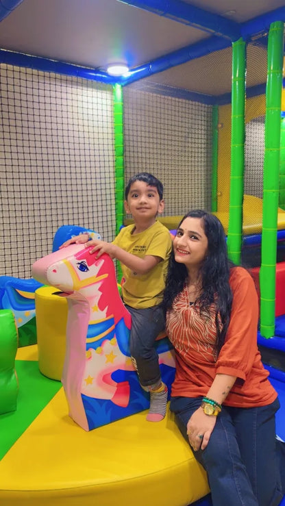 A woman and a young boy are playing in a colorful indoor playground. The boy is riding a pink unicorn toy.