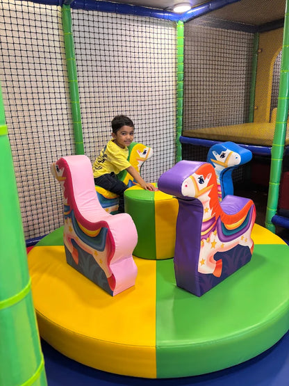 A young boy sits on a carousel with unicorn-themed seats in a colorful indoor playground.