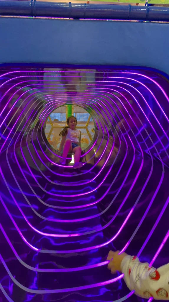 A young girl sits in a purple, illuminated tunnel at a play place.