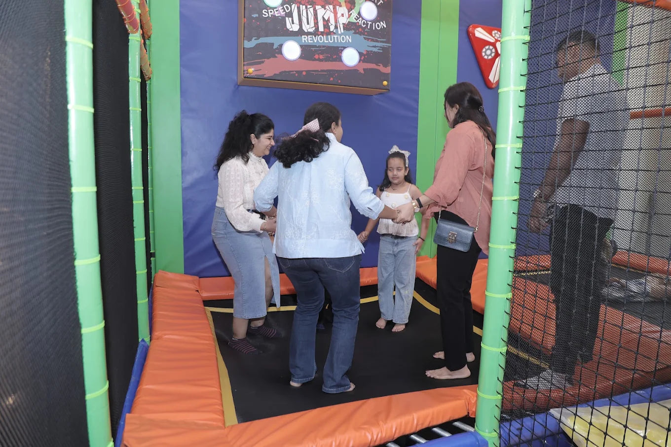 A group of adults and a child hold hands in a circle on a trampoline. They are in a colorful indoor play area with a "Jump Revolution" sign.