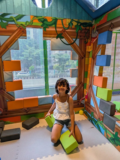 A young girl kneels on a padded floor in a colorful play area, holding large green blocks. She is surrounded by soft, oversized building blocks.