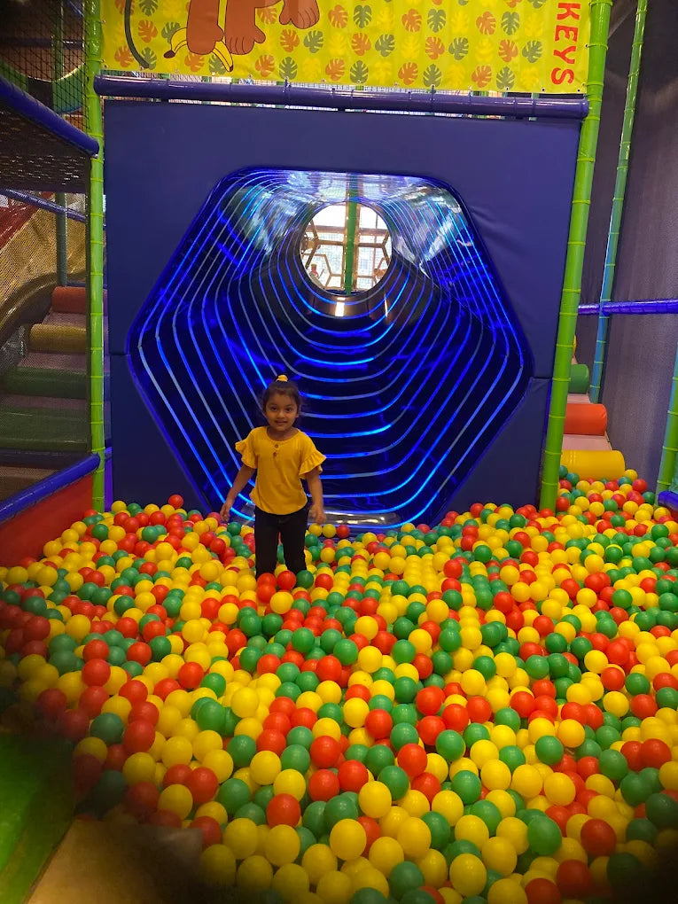 A young child stands in a ball pit in front of a blue, hexagonal tunnel with lights.