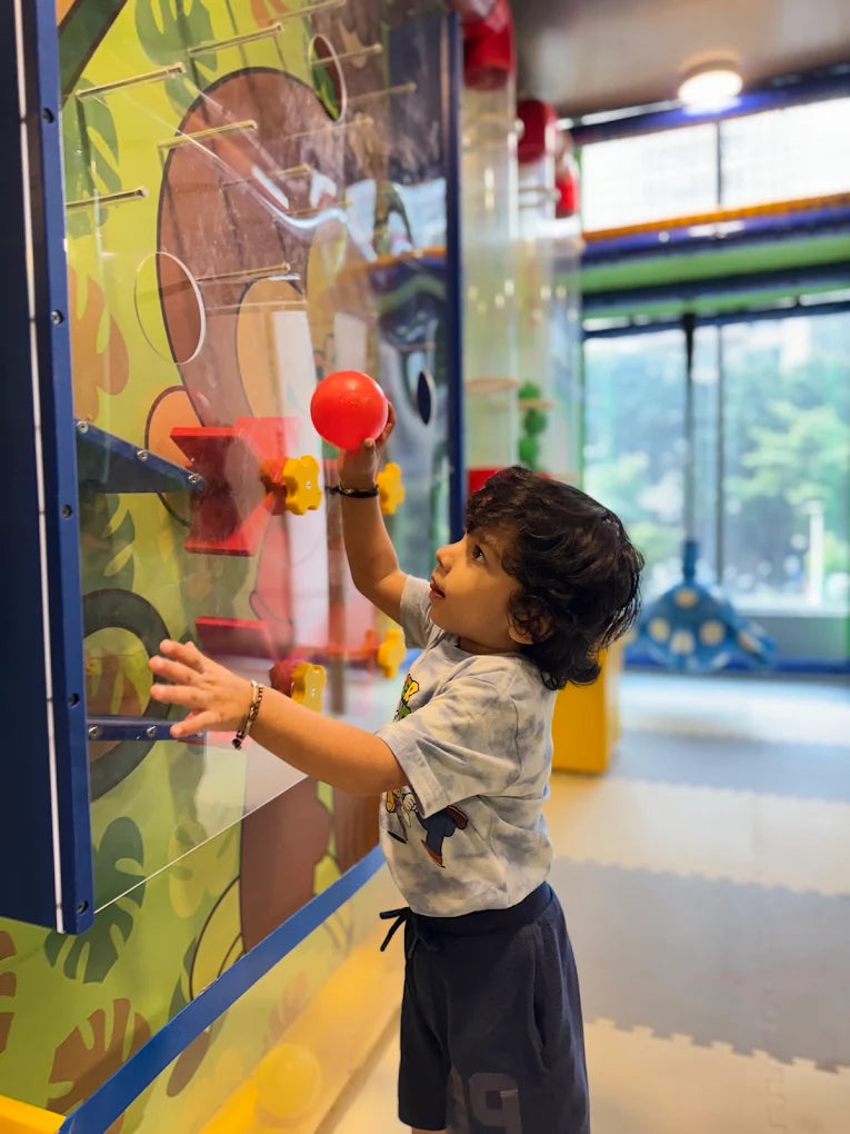 A young child plays with a red ball on a colorful interactive wall in a bright indoor play area.