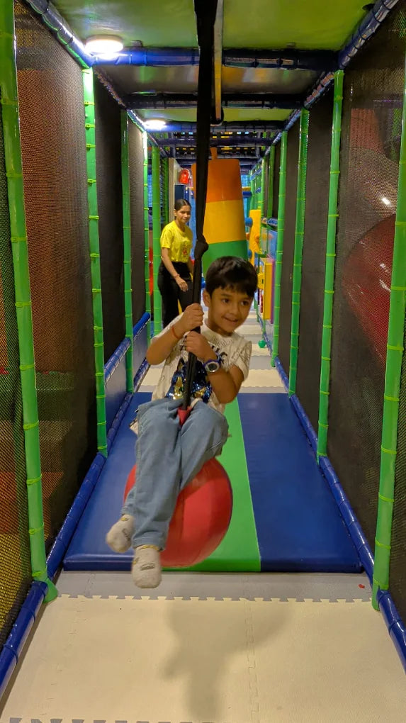 A young boy swings on a red ball in a colorful indoor playground. A woman stands in the background.