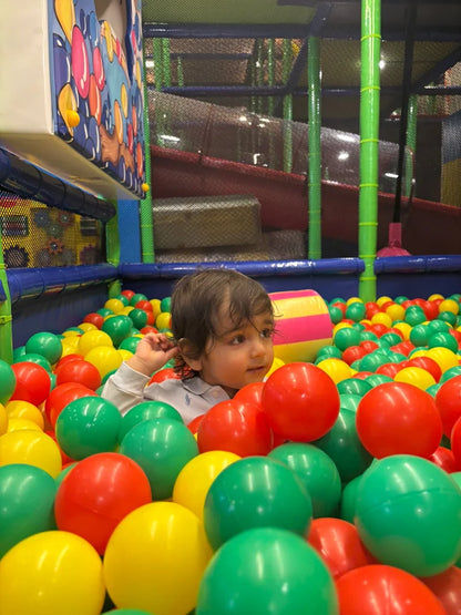 A young child plays in a ball pit filled with colorful balls. The child is surrounded by red, green, and yellow balls.