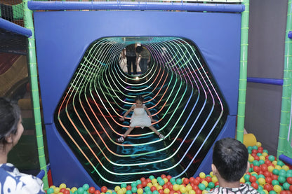 A young girl slides down a colorful, illuminated tunnel in a play area, with other children and adults nearby.