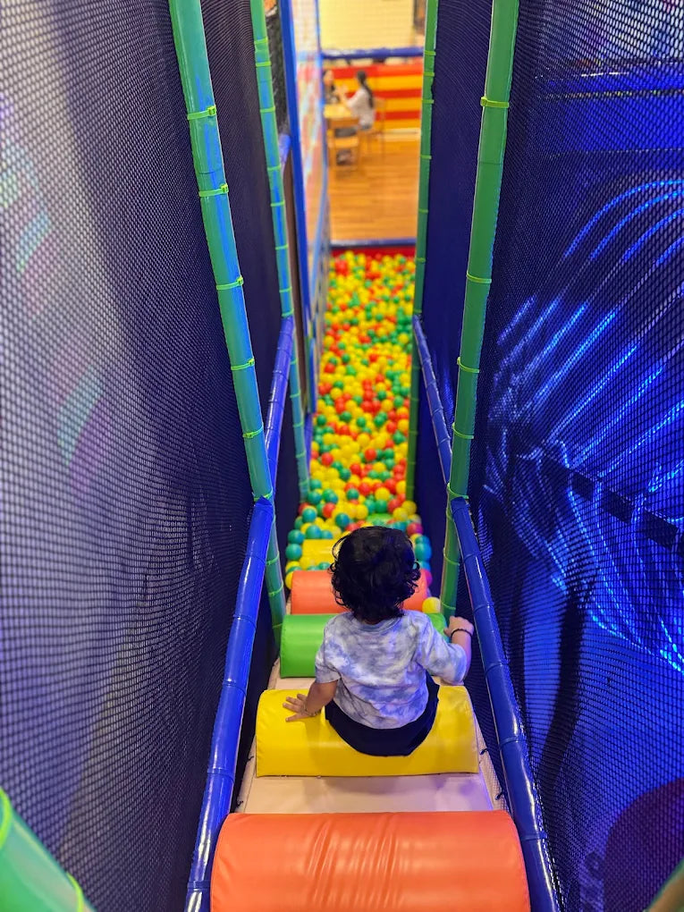 A young child sits on a yellow mat in a ball pit play area, looking down at the colorful balls.