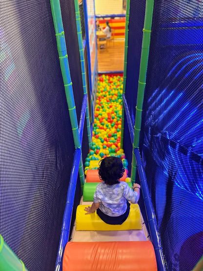 A young child sits on a yellow mat in a ball pit play area, looking down at the colorful balls.