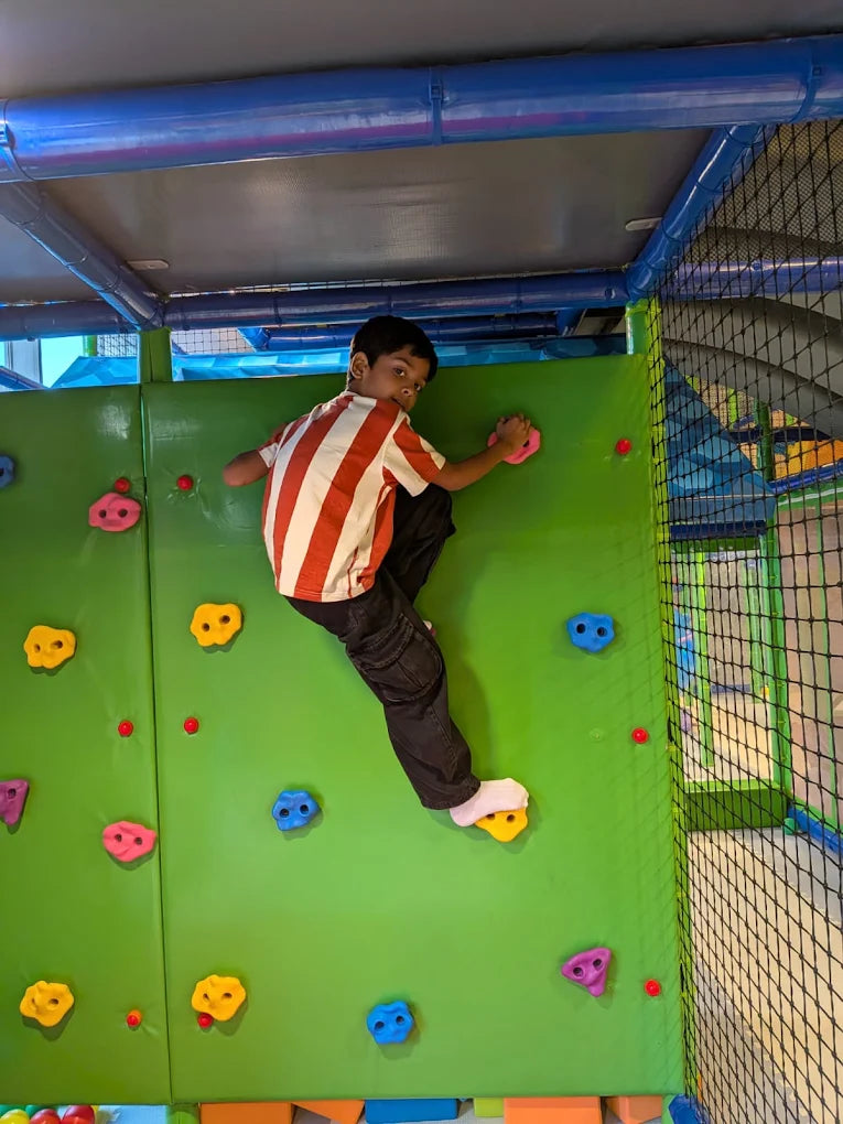 A young boy climbs a green wall with colorful holds in an indoor play area.