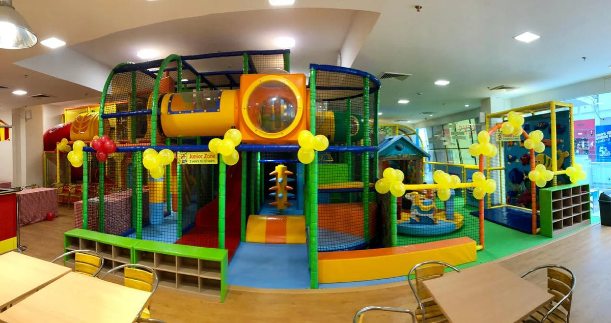 A colorful indoor playground with slides, tunnels, and ball pits for young children. Tables and chairs are set up in the foreground.