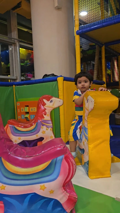 A young child stands in a colorful indoor playground next to a yellow soft play structure.