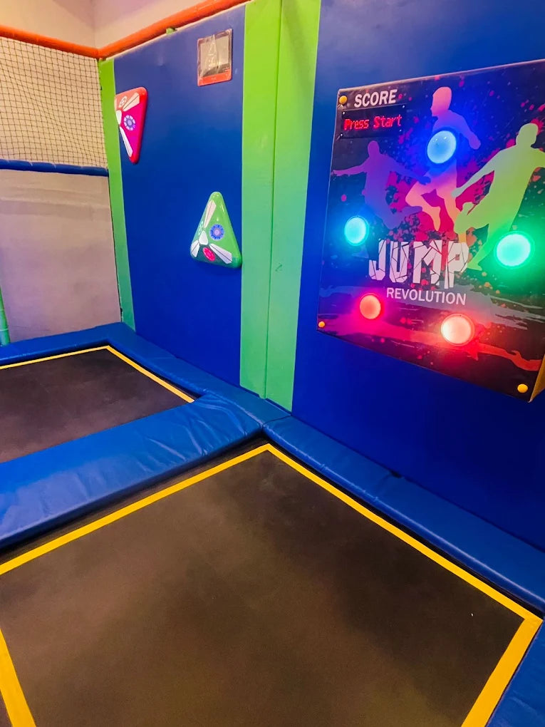 A trampoline park with colorful walls, climbing holds, and a "Jump Revolution" game screen.