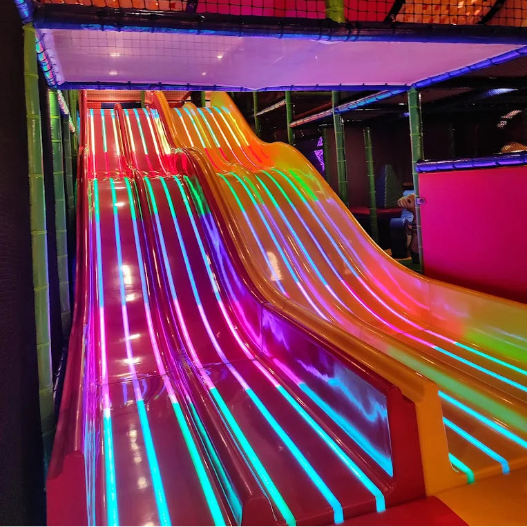 A brightly lit indoor playground with colorful slides. Children are playing on the slides.