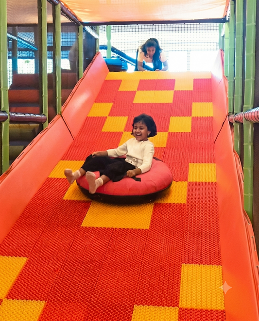 A young child slides down a colorful indoor playground slide on a tube, while an older child watches from the top.