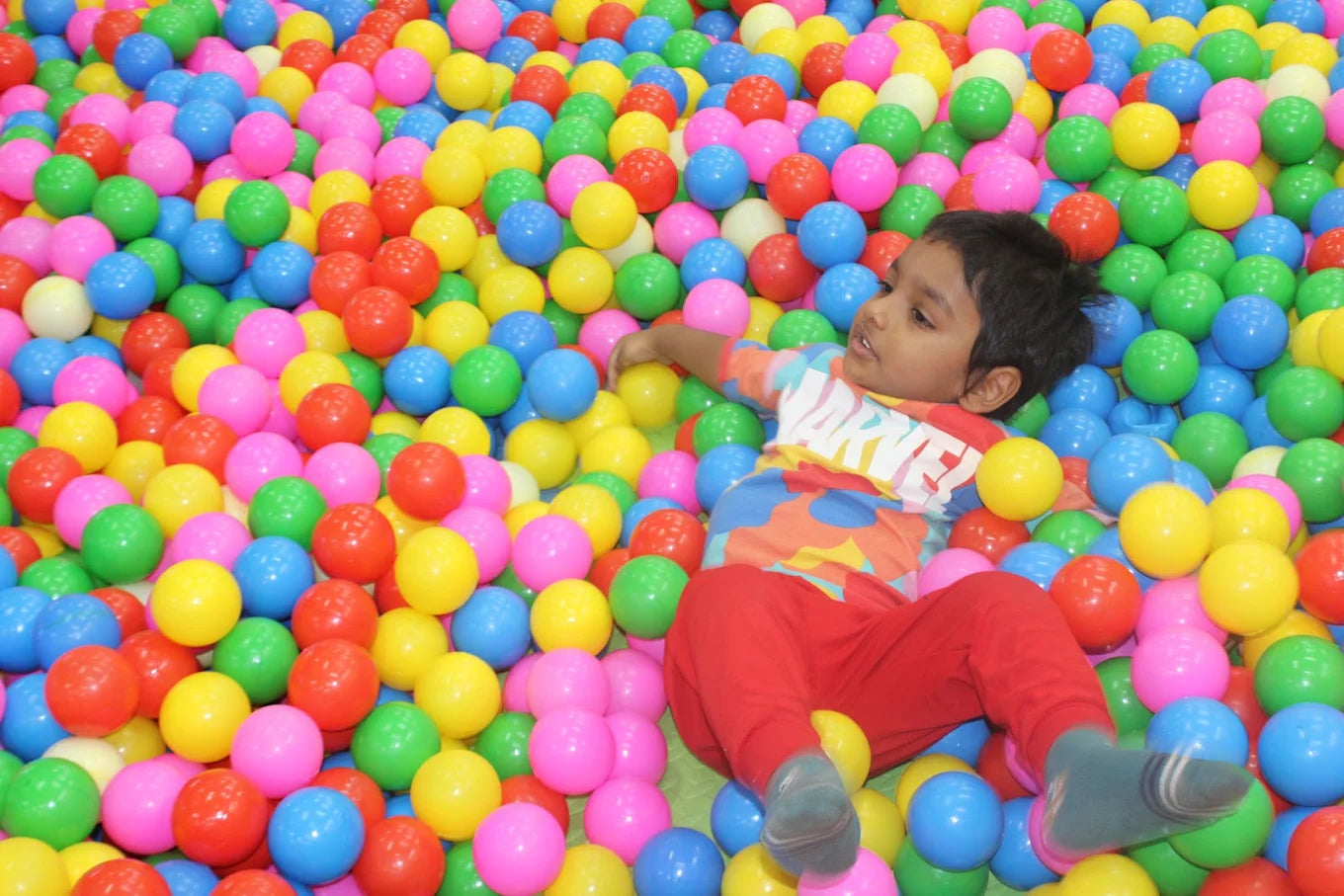 A young child lies in a ball pit, surrounded by colorful balls.