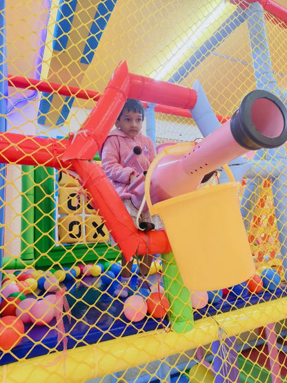 A young child in a pink jacket sits in a colorful indoor playground with a yellow bucket.
