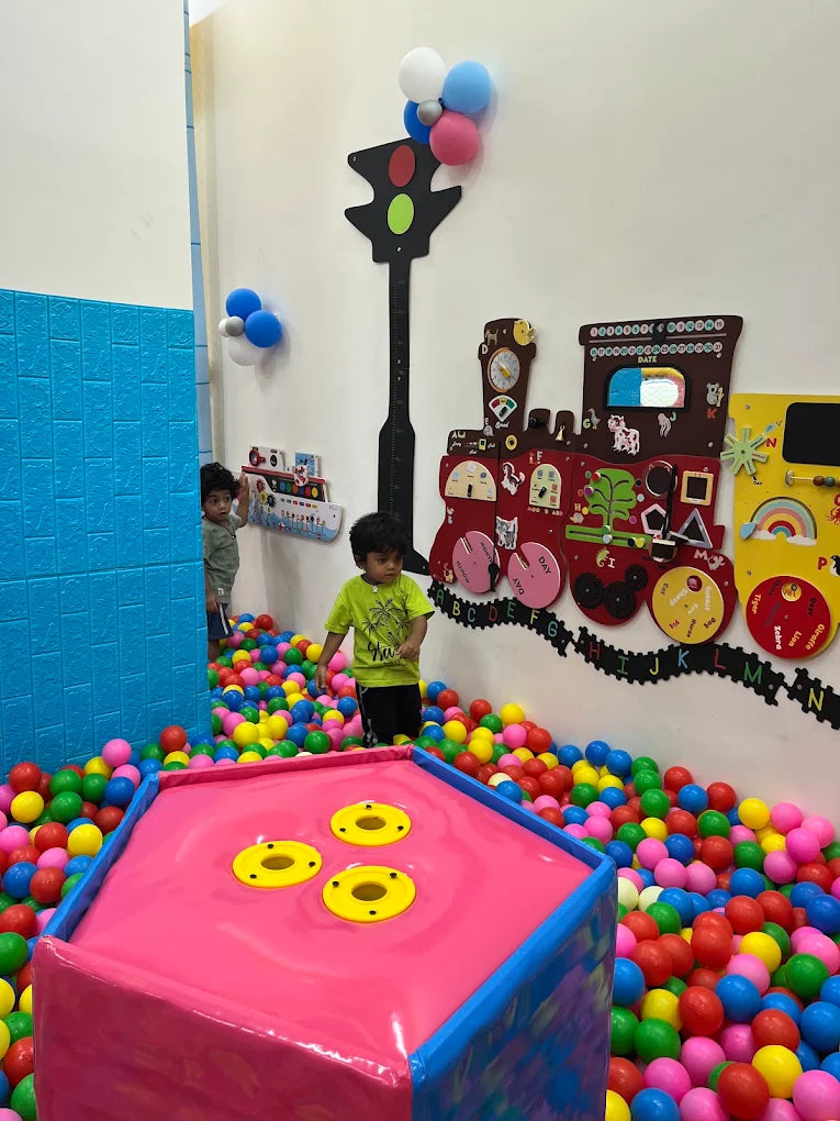 Children play in a ball pit with a train-themed wall decoration.
