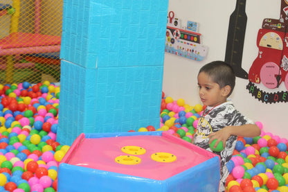 A young boy plays in a ball pit at an indoor playground. He holds a green ball, looking at a pink and blue game table.