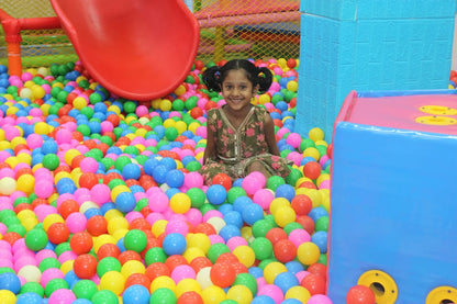 A young girl smiles while sitting in a ball pit with a red slide in the background.
