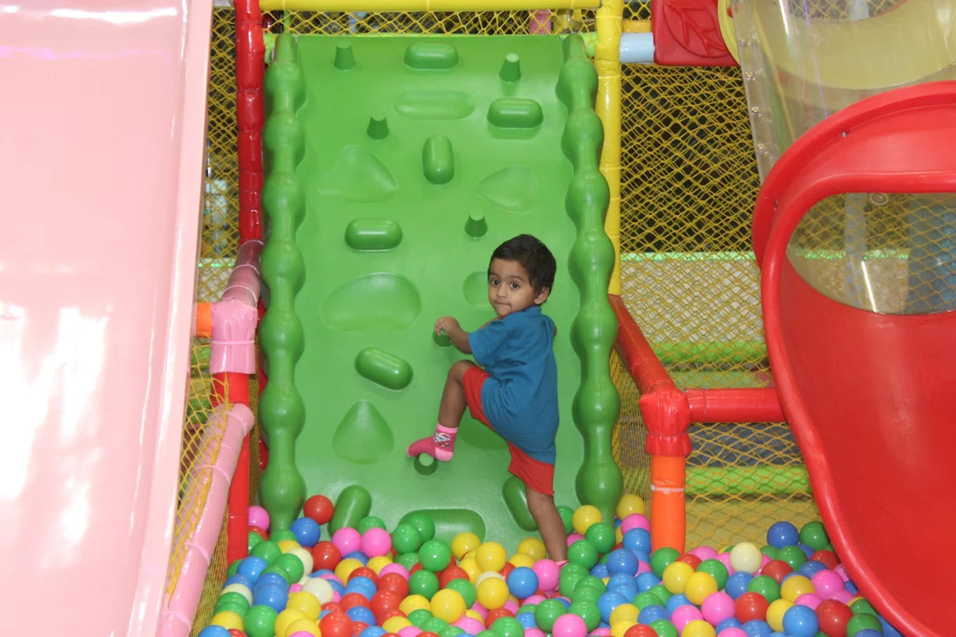 A young child climbs a green wall in a ball pit at a playground.