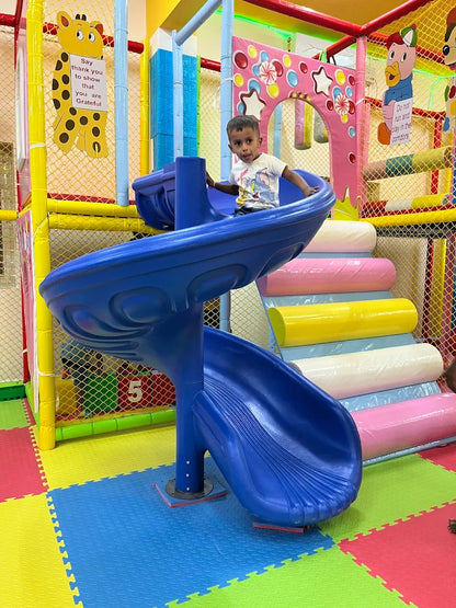 A young boy sits on a blue slide in a colorful indoor playground. He is looking at the camera with a slight smile.