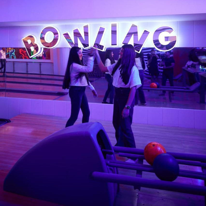 Two people at a bowling alley with a neon 'Bowling' sign in the background.
