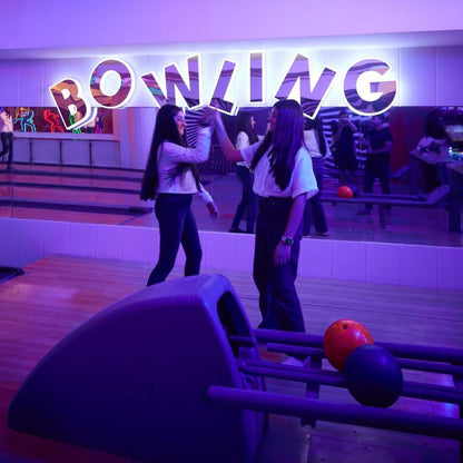 Two people at a bowling alley with a neon 'Bowling' sign in the background.