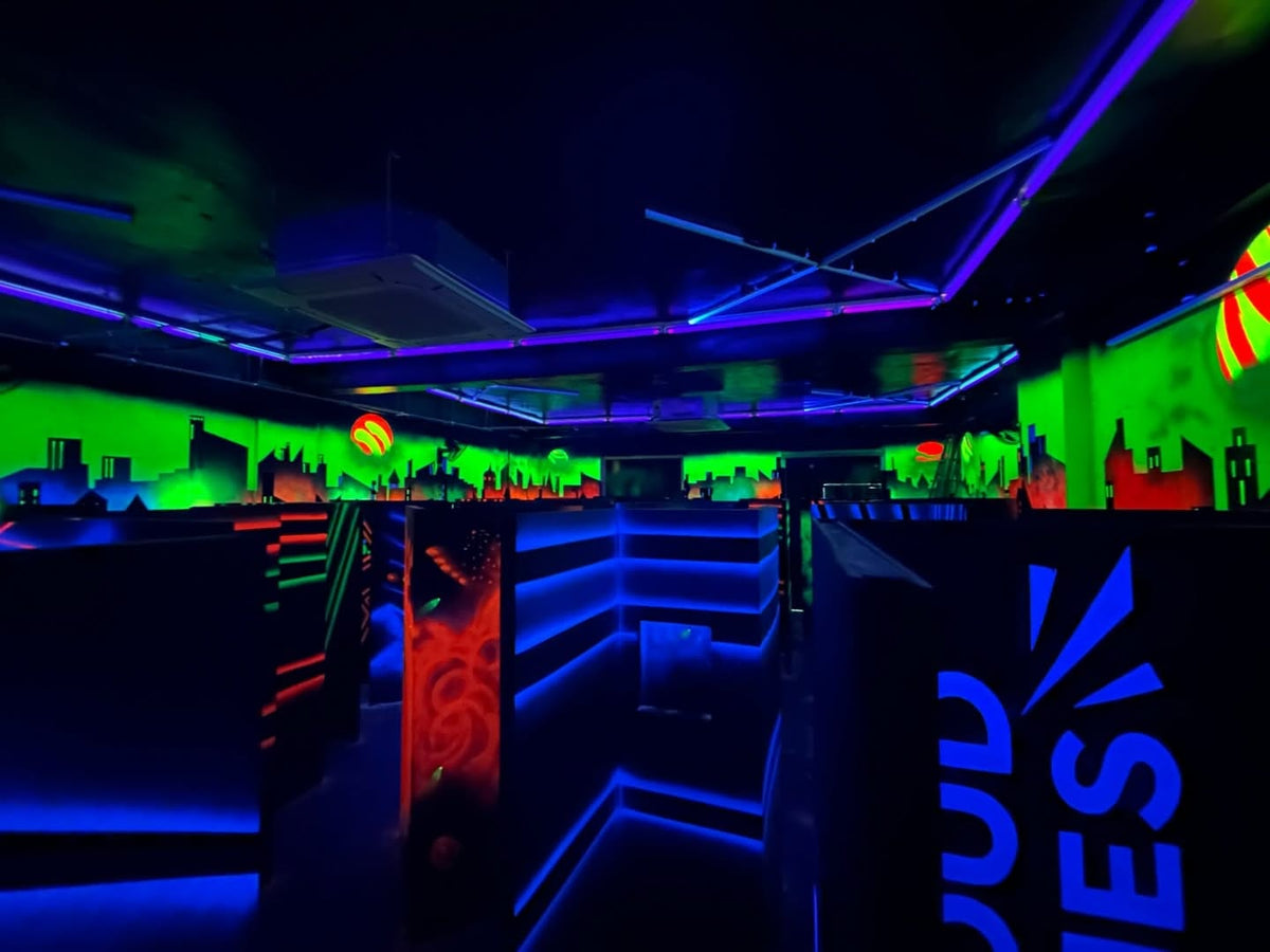 A neon-lit laser tag arena with glowing blue and green walls, dark maze-like structures, and a cityscape mural.